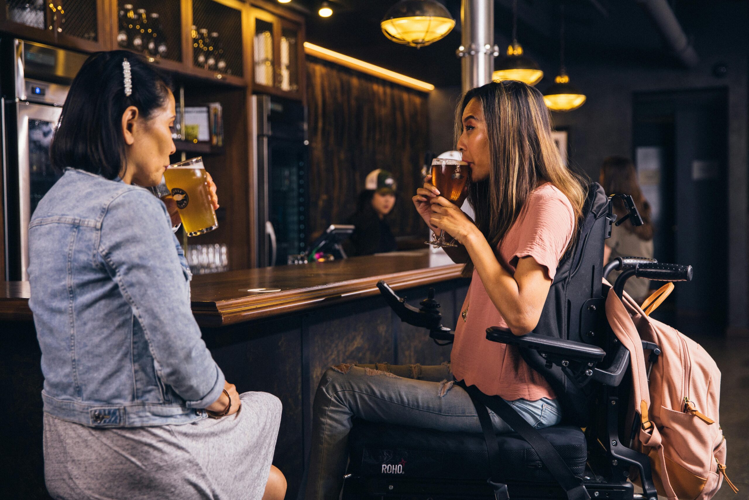 Two women enjoying beer at a bar, one seated in a wheelchair, capturing a moment of camaraderie.